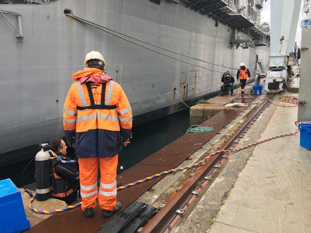 Trabajadores junto a un gran barco gris, uno con equipo de seguridad naranja, otros cerca.