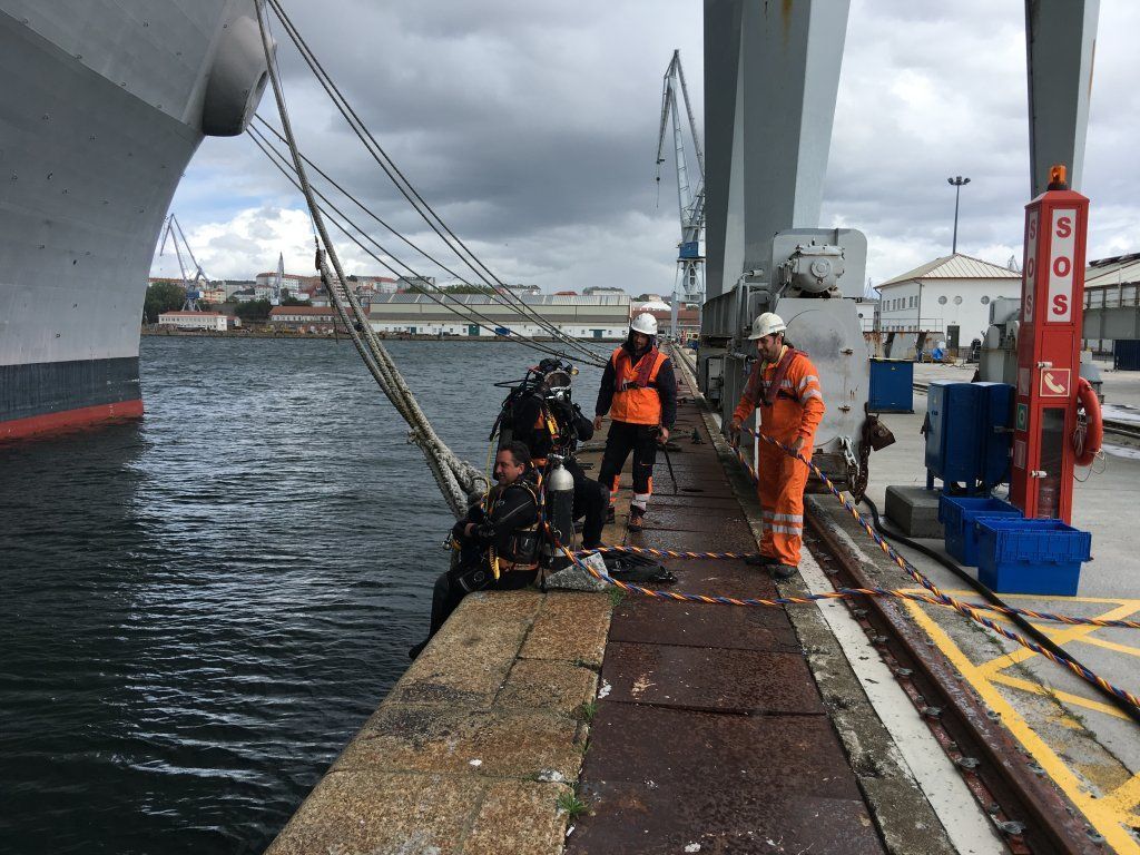 Buzos preparándose para trabajos submarinos cerca de un gran buque en un puerto. Personas con equipo de seguridad en un muelle.