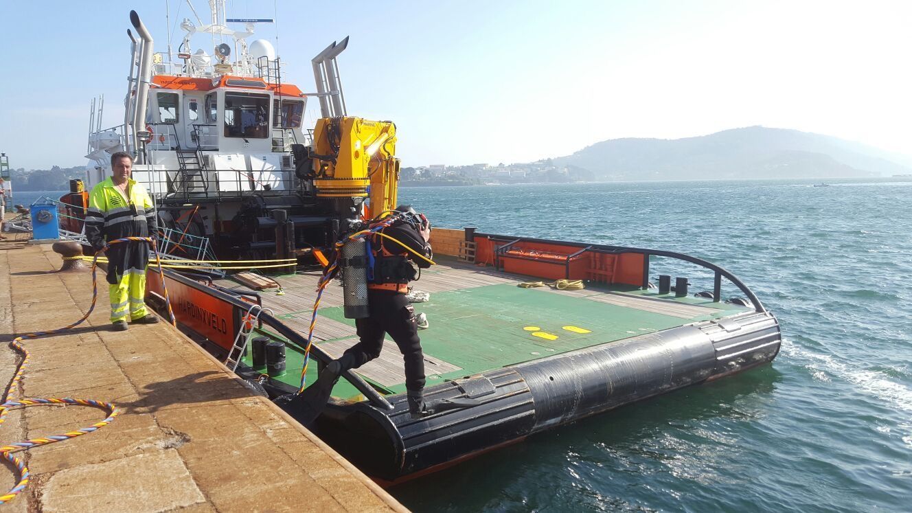 Buzo saliendo del agua a un barco de trabajo junto a un muelle. El barco tiene maquinaria y hay otra persona cerca. Día soleado con el mar al fondo.