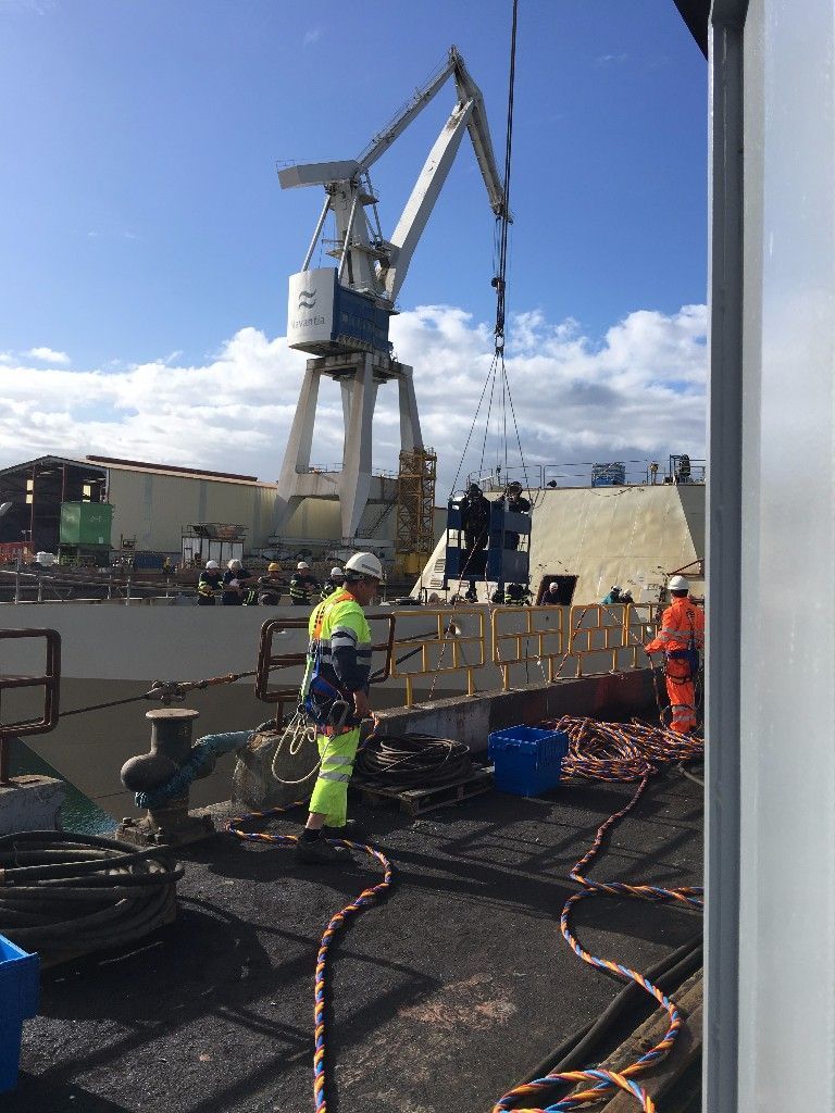 Trabajadores cerca de una gran grúa en una obra. Un trabajador con equipo de seguridad está de pie en una cubierta, con el cielo azul por encima.