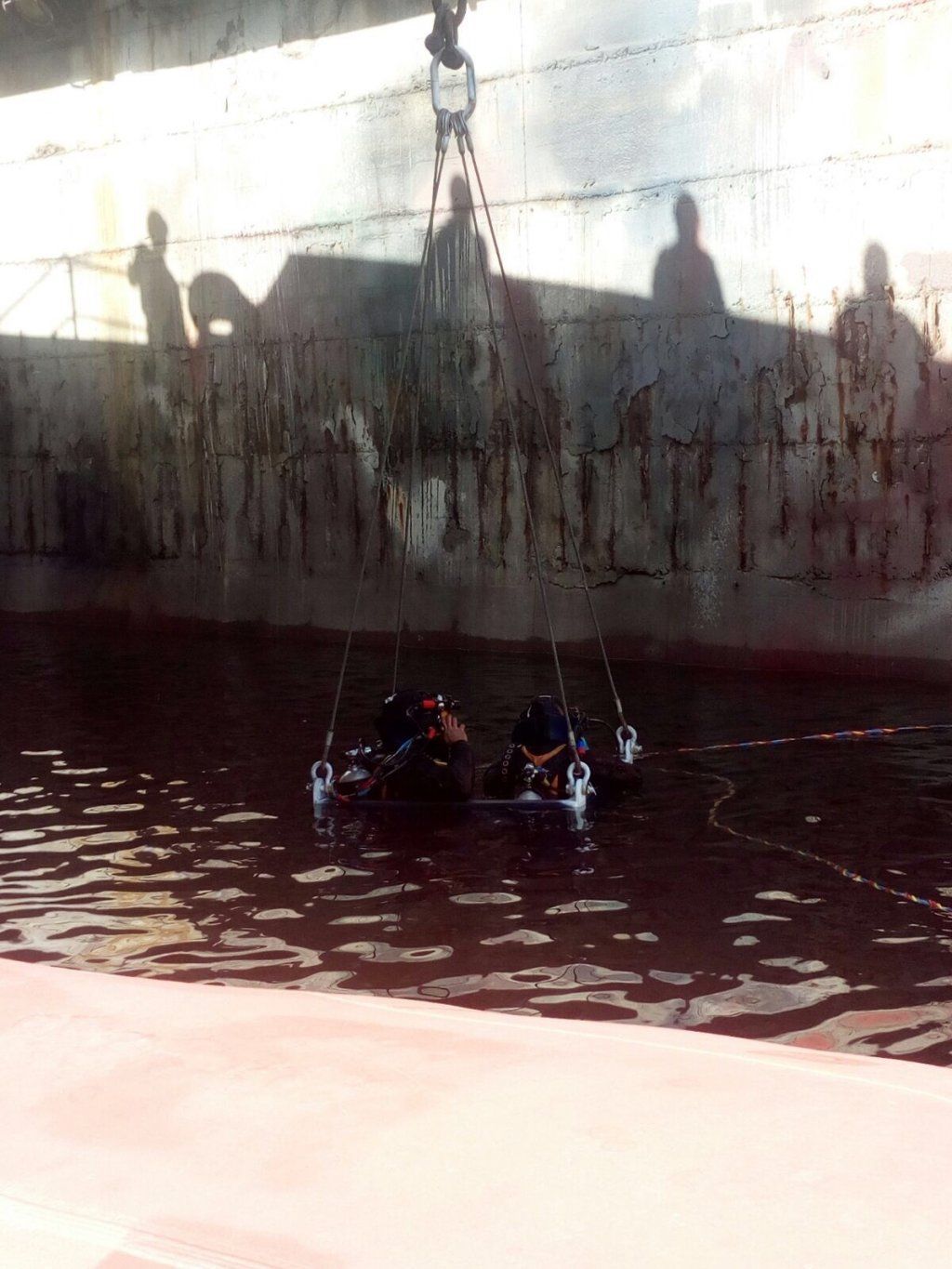 Buzos en el agua bajo un gran barco, mientras una grúa lo baja. Se ven las sombras de los trabajadores en el cielo.