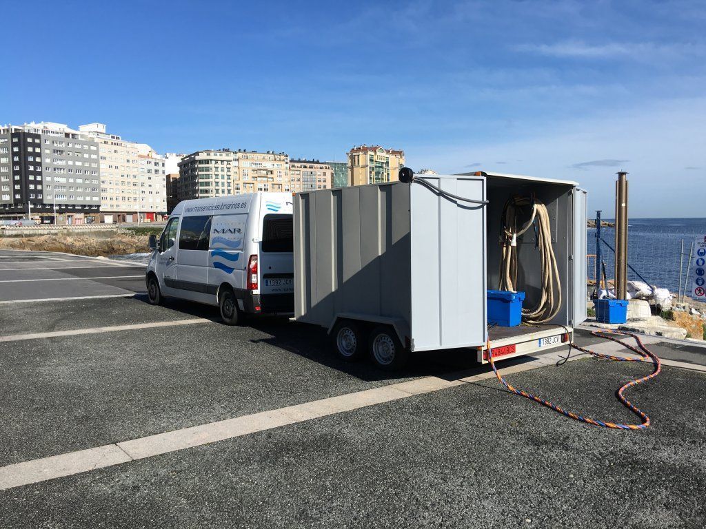 Una furgoneta arrastra un remolque cerca de la costa; cielo azul y edificios al fondo. El remolque tiene una manguera conectada.