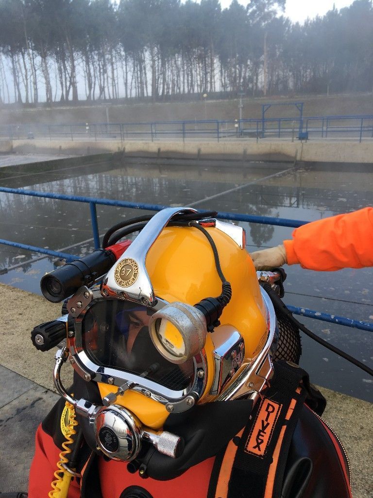 Buzo con casco amarillo, cerca de un cuerpo de agua y con equipo de buceo. Una persona con manga naranja le ayuda.