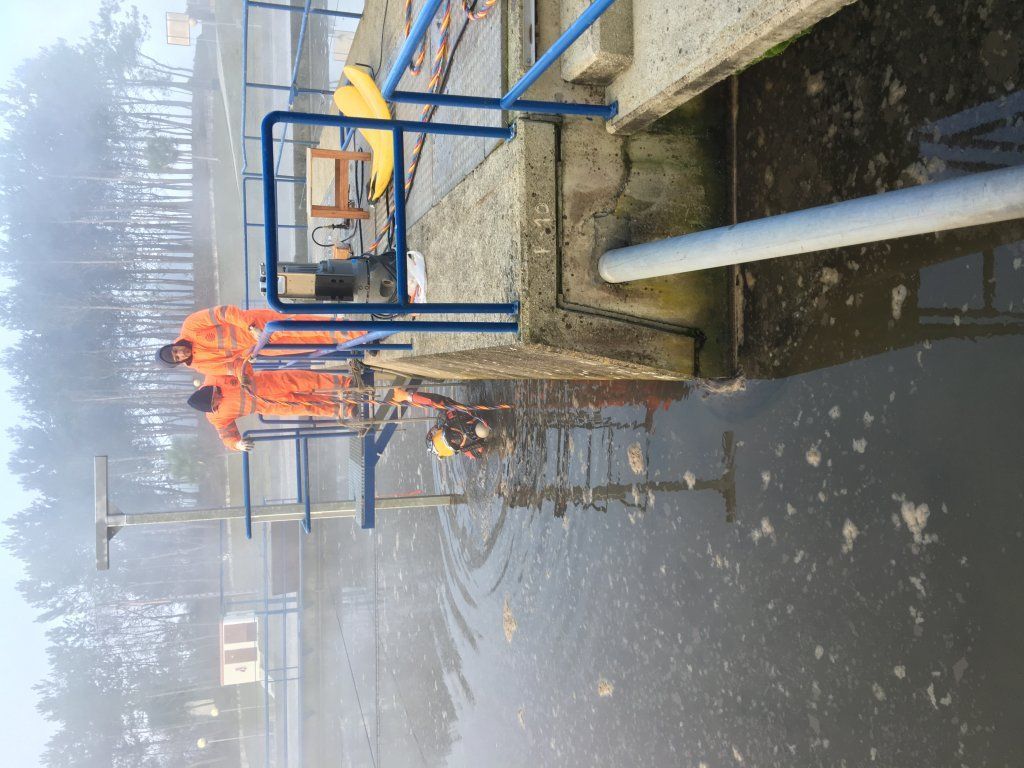 Dos trabajadores con trajes protectores de color naranja cerca del agua, cerca de una estructura de hormigón, en el exterior con niebla.