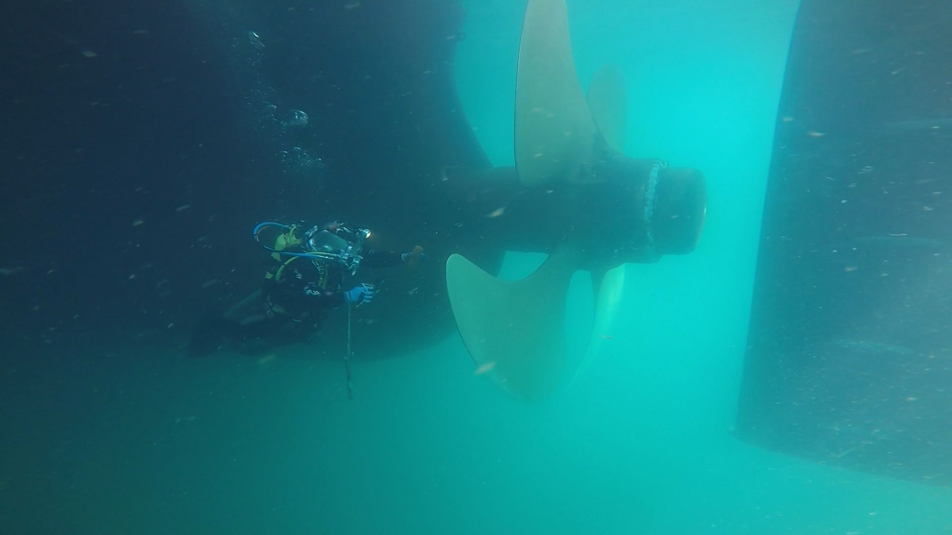Buzo inspeccionando la hélice de un gran barco bajo el agua, cerca de un casco oscuro. Aguas turquesas.
