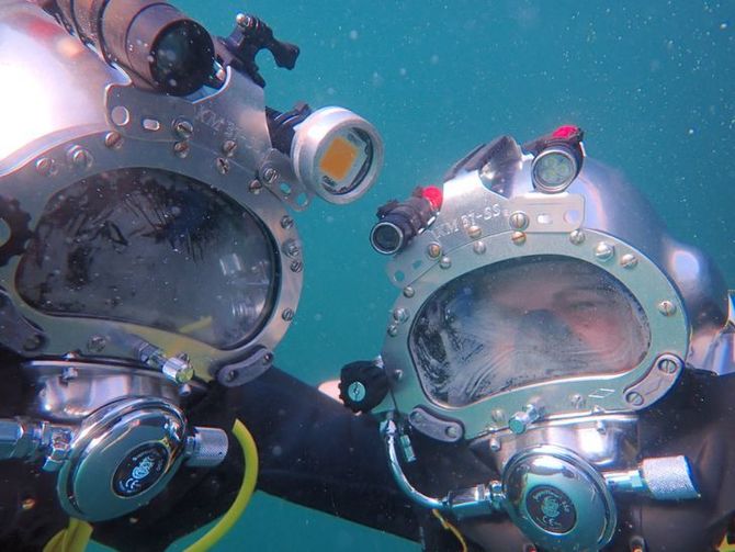 Dos buceadores que llevan cascos de buceo plateados vintage bajo el agua, uno frente al otro.
