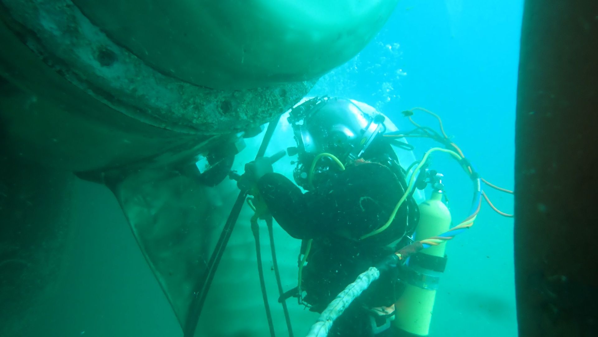 Buzo con traje negro inspeccionando un barco bajo el agua. Agua verde.