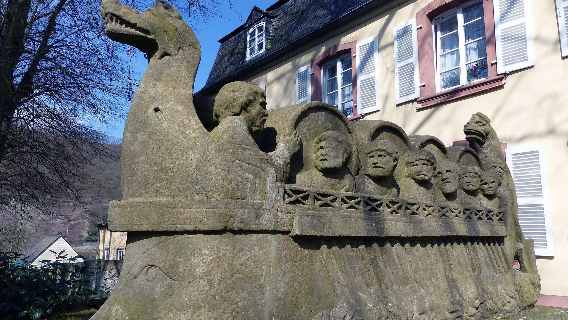 Steinskulptur eines Wikingerschiffs mit eingemeißelten Gesichtern vor einem Gebäude im Sonnenlicht
