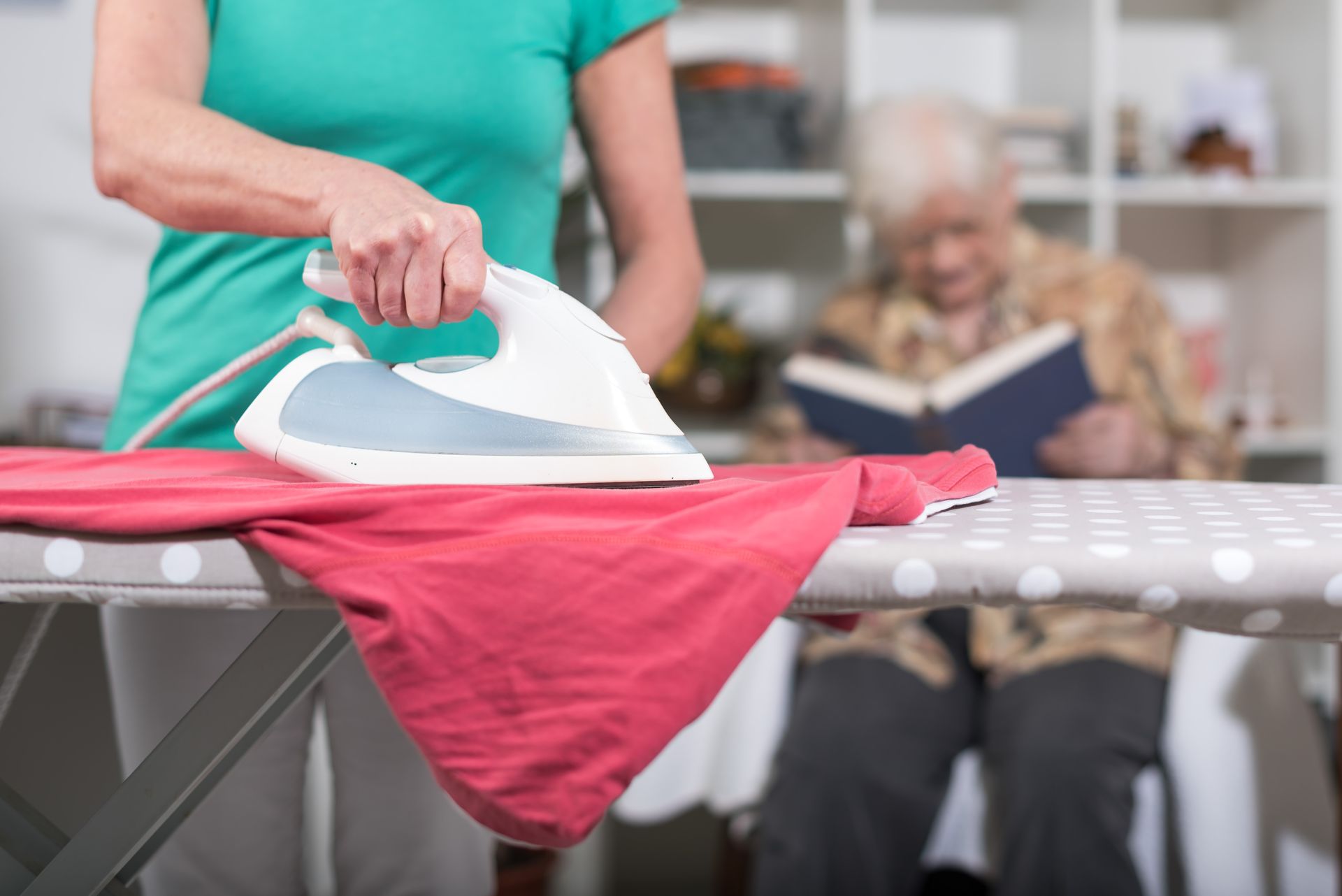 Une personne repasse une chemise rose sur une planche à repasser tandis qu'une personne âgée lit en arrière-plan