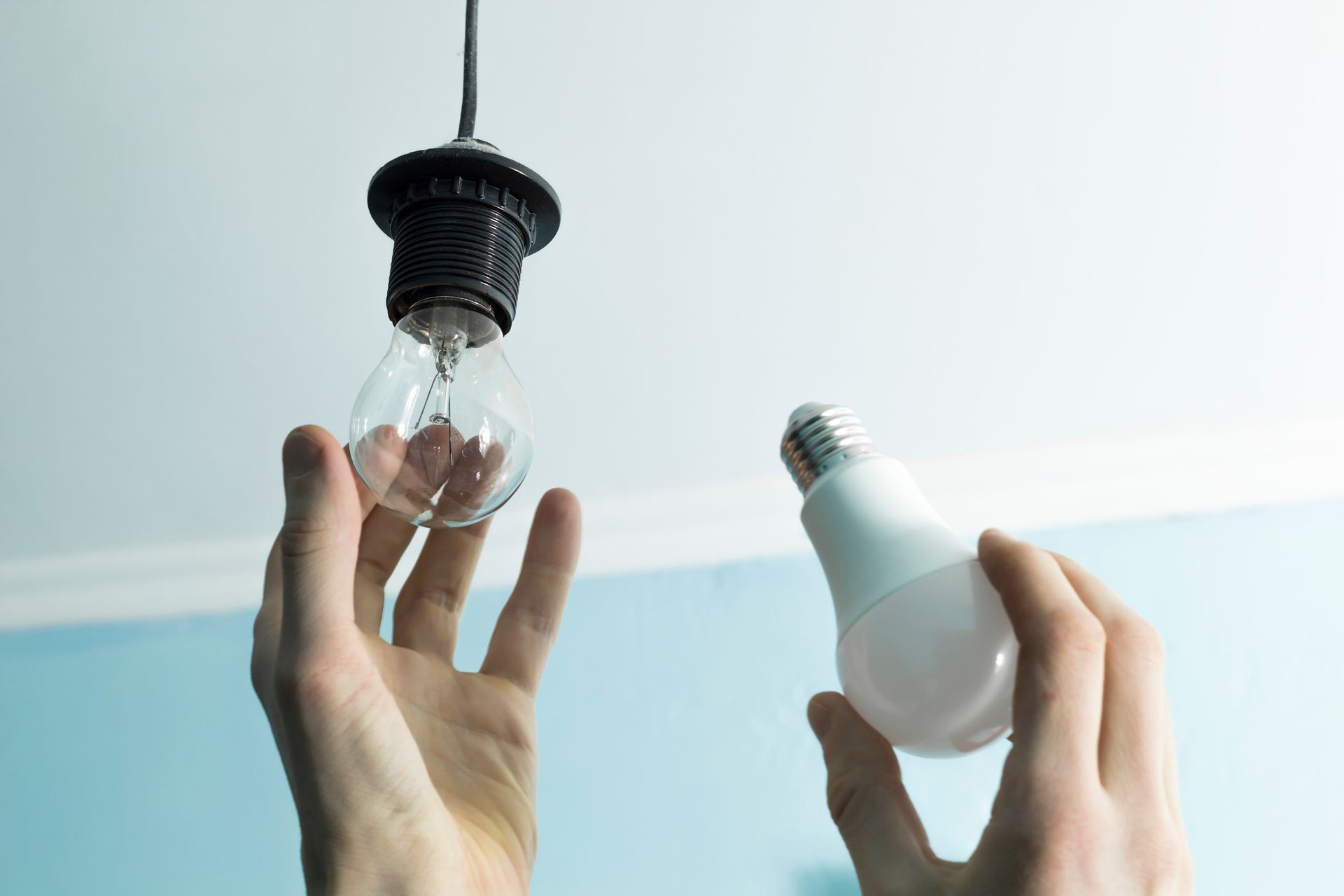 Des mains tenant une ampoule transparente cassée et une ampoule blanche intacte contre un mur bleu pâle