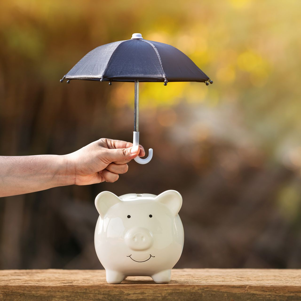 Une main tenant un minuscule parapluie au-dessus d'une tirelire blanche posée sur une surface en bois