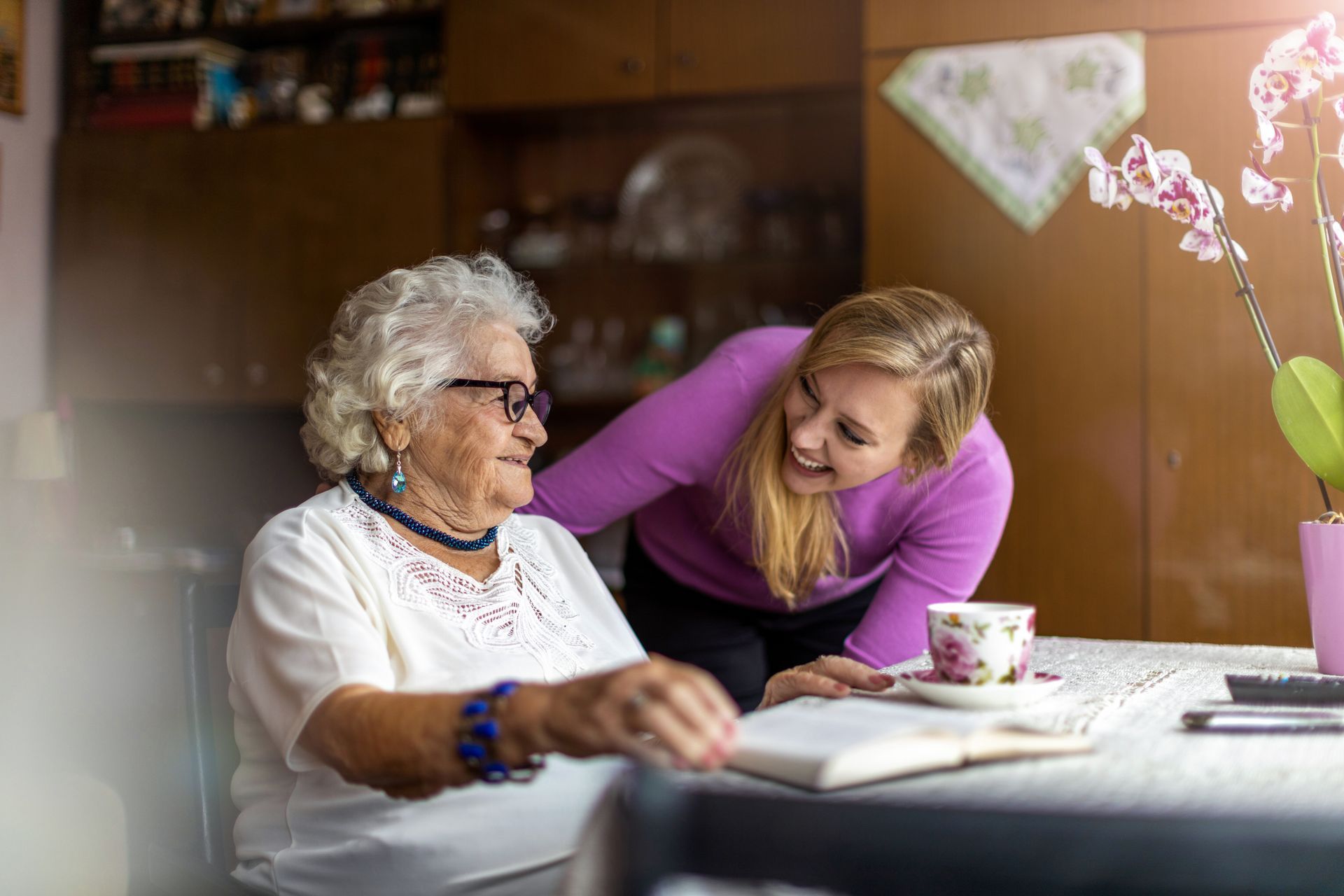 Une femme se penche vers une femme âgée assise à une table, avec qui elle échange une conversation