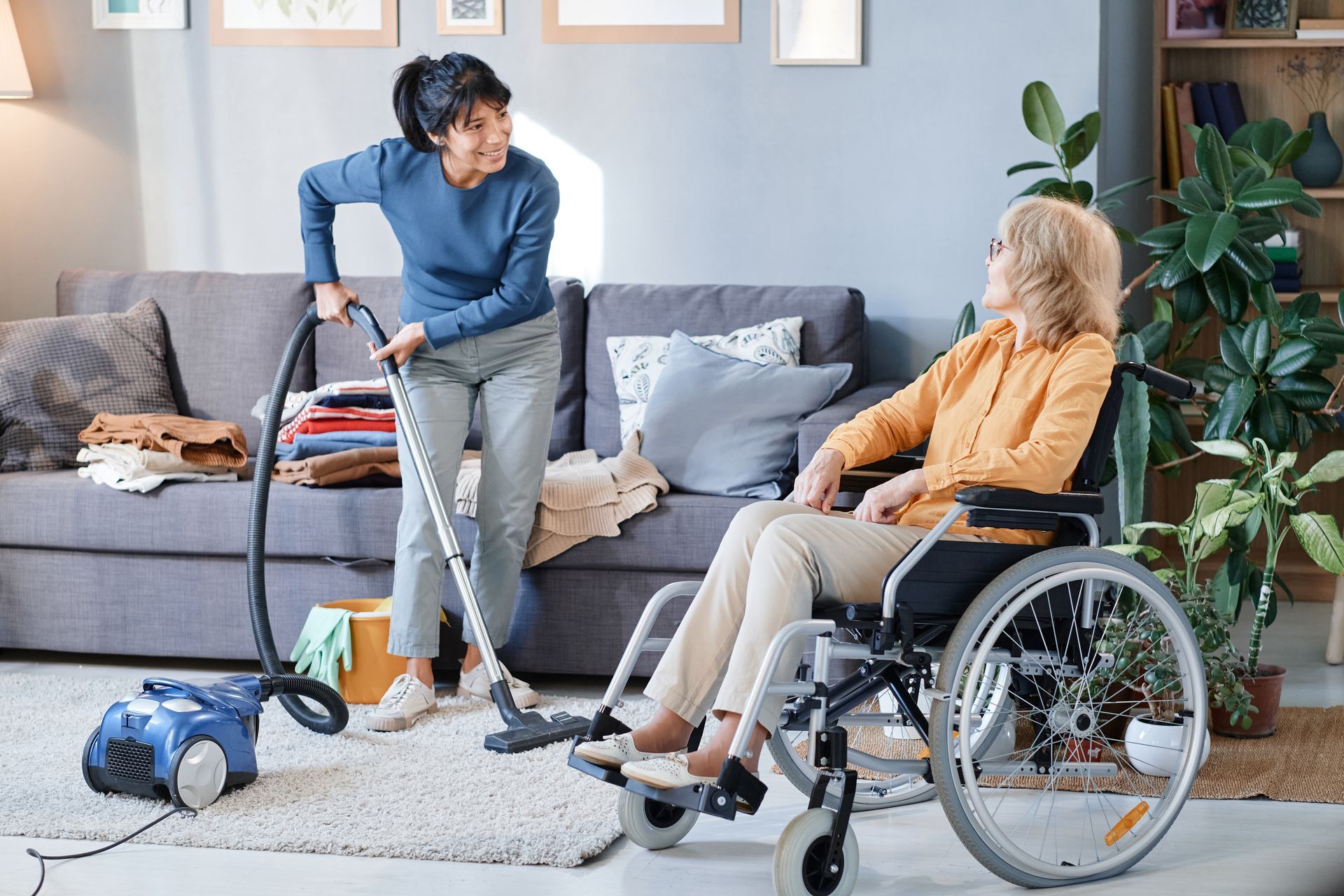 Une personne passe l'aspirateur dans un salon tandis qu'une personne en fauteuil roulant est assise à proximité sur un canapé gris