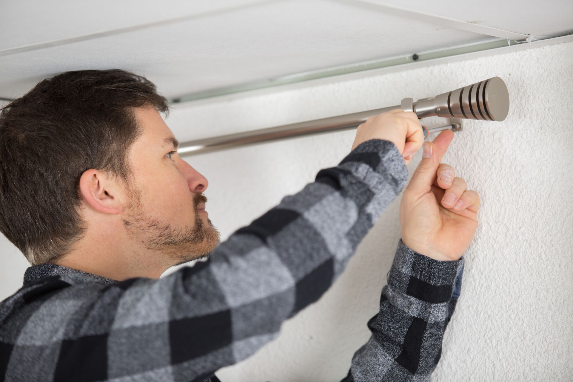 Une personne installe une tringle à rideaux sur un mur blanc, tenant une perceuse d'une main et la tringle de l'autre