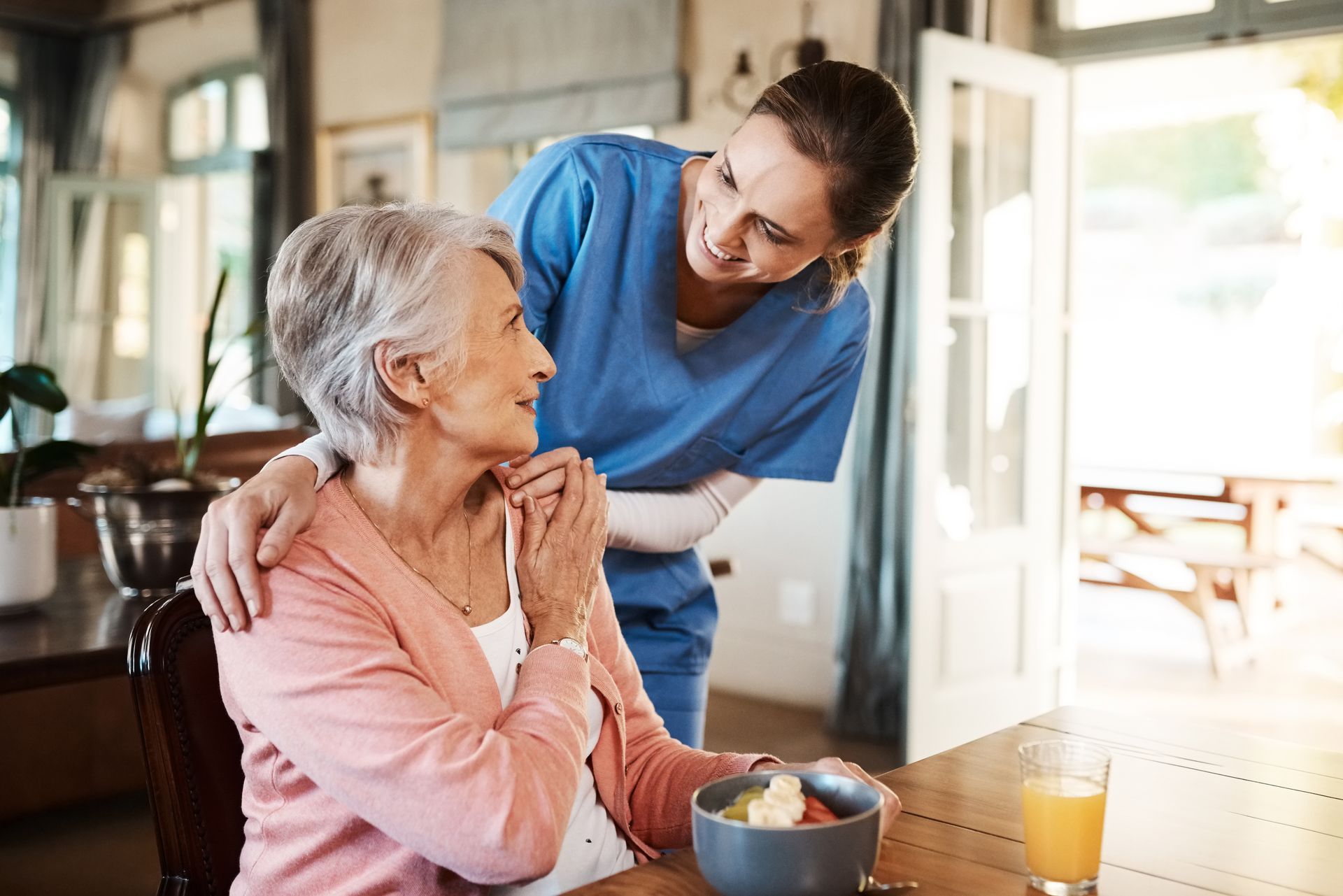 Une aide-soignante se penche en souriant à une femme âgée assise à une table à manger dans une cuisine