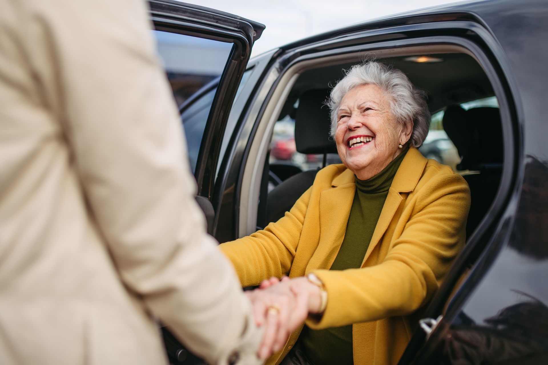 Une femme souriante, vêtue d'un cardigan moutarde, sourit à une personne l'aidant à sortir de la voiture
