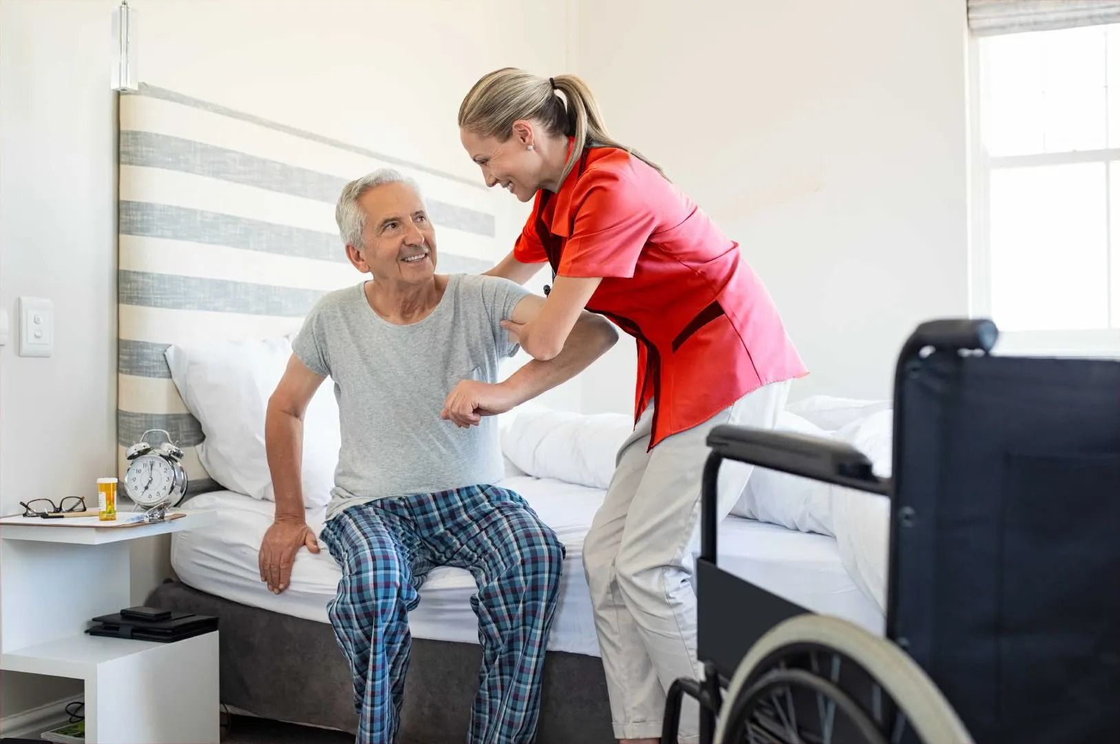 Une aide-soignante aide un patient à s'asseoir sur un lit dans une chambre, un fauteuil roulant se trouvant à proximité