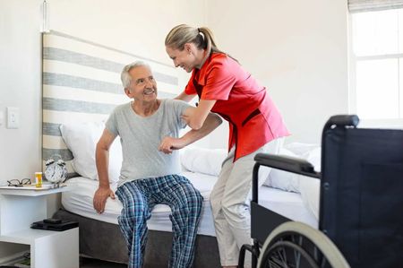 Une aide-soignante aide un patient à s'asseoir sur un lit dans une chambre, un fauteuil roulant se trouvant à proximité