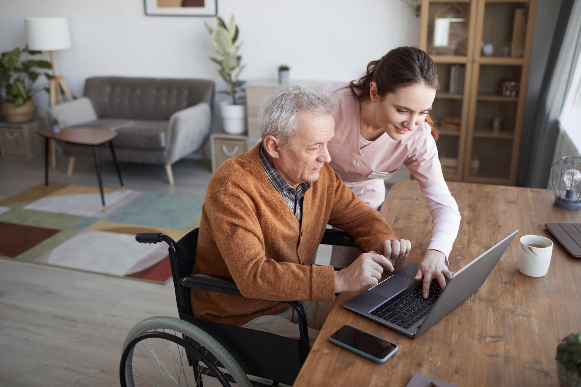 Une aide-soignante aide un homme en fauteuil roulant à utiliser un ordinateur portable à une table en bois dans un salon