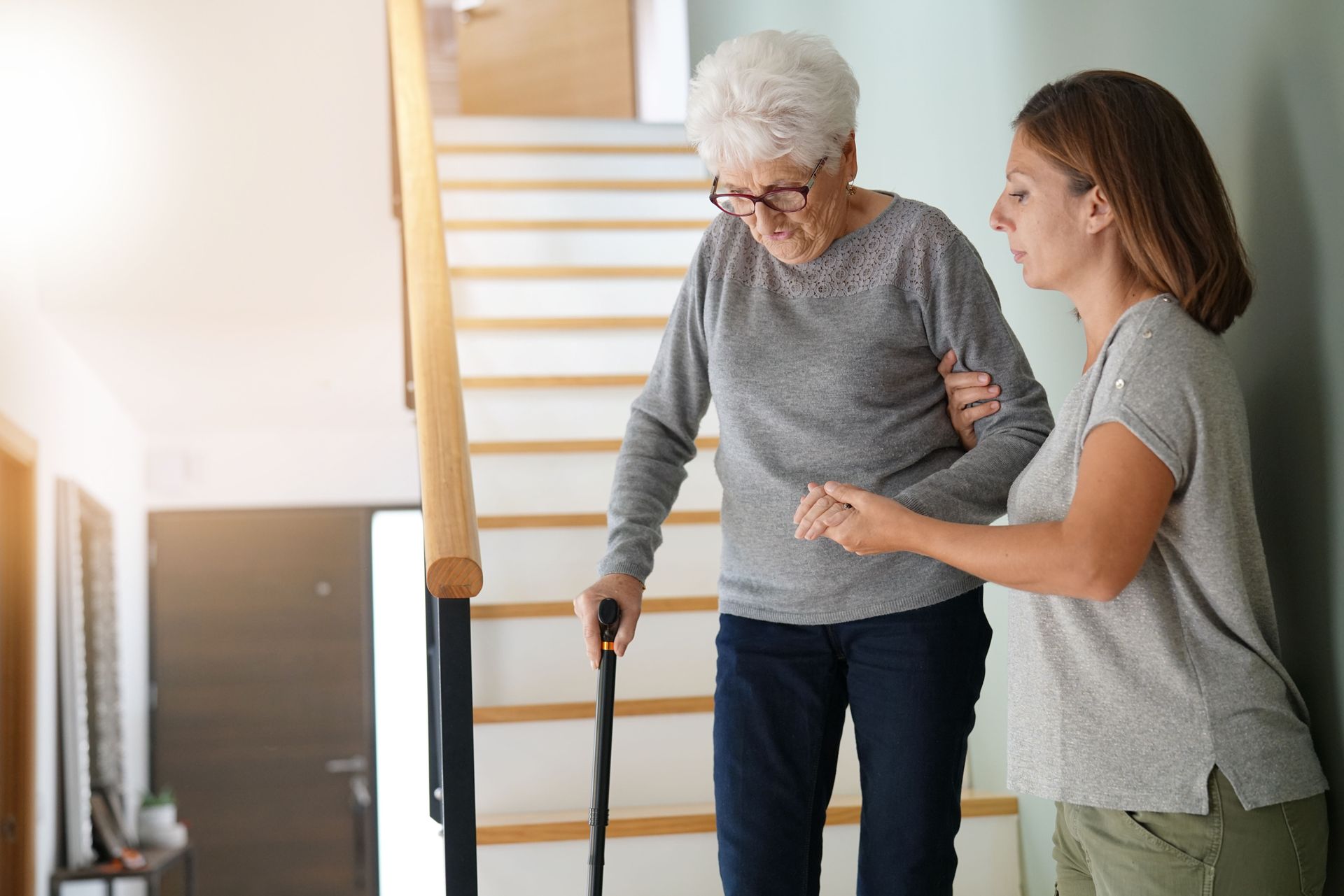 Une femme aide une personne âgée avec une canne près d'un escalier dans un intérieur lumineux
