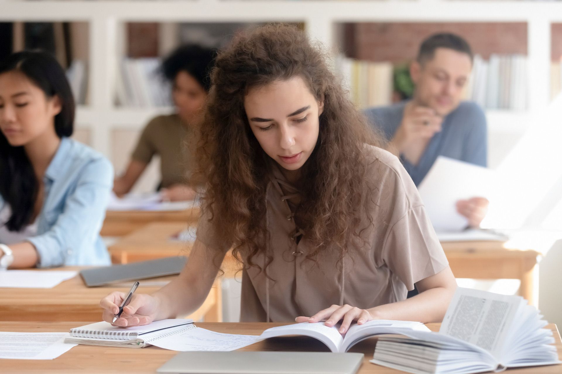 Estudiantes estudiando en un aula; una mujer escribe en un cuaderno mientras otros leen libros.