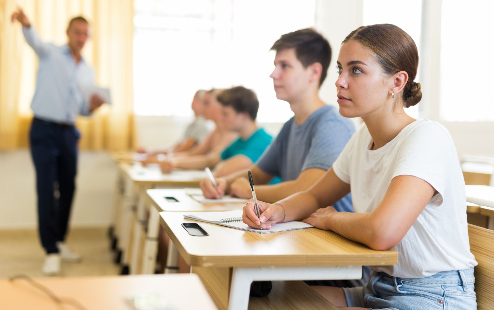 Alumnos realizando un examen en clase mientras un profesor permanece de pie al frente.