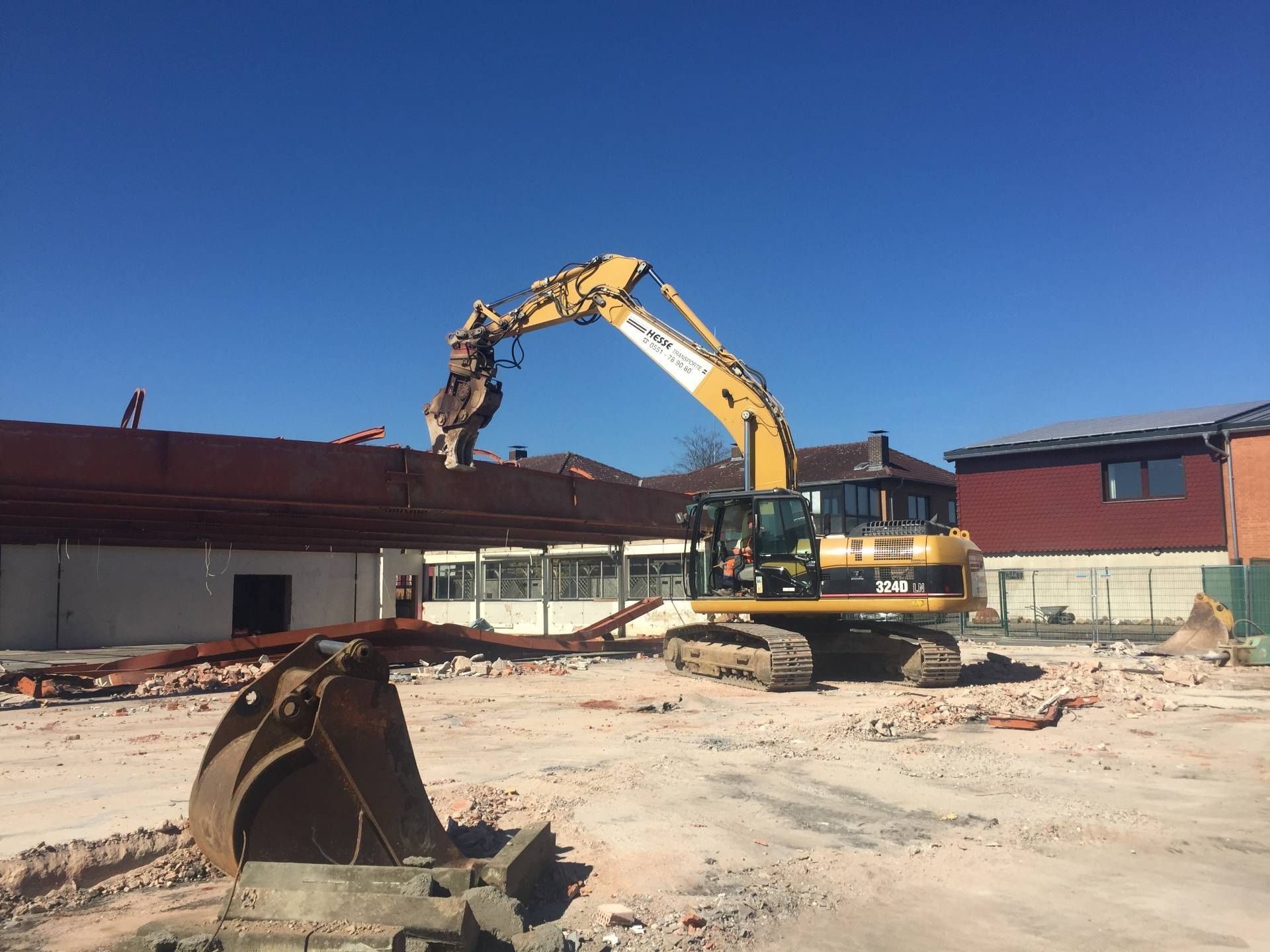 Ein gelber Bagger reißt unter klarem blauen Himmel ein niedriges rotes Gebäude auf einer Baustelle ab.