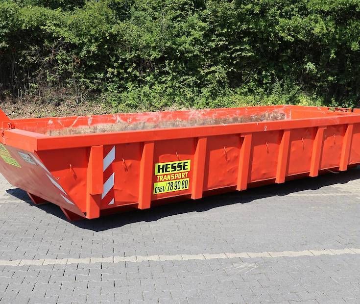 Ein leuchtend orangefarbener Bauschuttcontainer steht am Straßenrand neben grünen Büschen.
