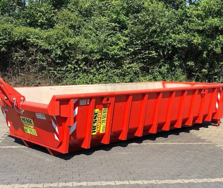 Ein roter Bauschuttcontainer steht auf dem Gehweg neben grünen Büschen.