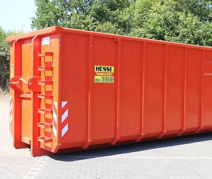Ein orangefarbener Industriecontainer steht im Freien auf einer gepflasterten Fläche; im Hintergrund sind Bäume zu sehen.