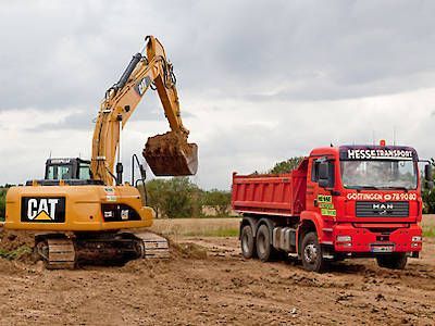 Ein gelber CAT-Bagger lädt Erde in einen roten Hesserbäçk-Muldenkipper auf einer schlammigen Baustelle.