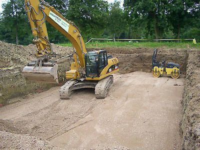 Ein gelber Bagger und eine kleine Walze verdichten einen Feldweg auf einer Baustelle.