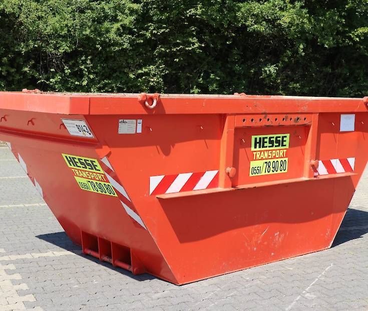 Ein roter Bauschuttcontainer mit Warnaufklebern steht auf einer gepflasterten Straße vor grünen Bäumen.
