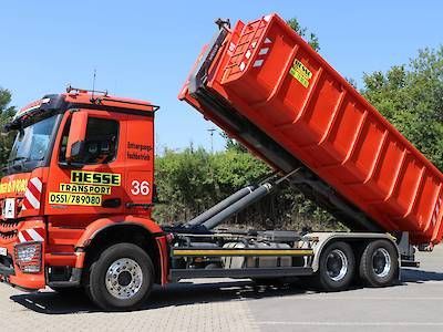 Ein roter Abschleppwagen mit Pritsche hebt einen umgekippten roten Müllcontainer auf einer Straße unter klarem Himmel an