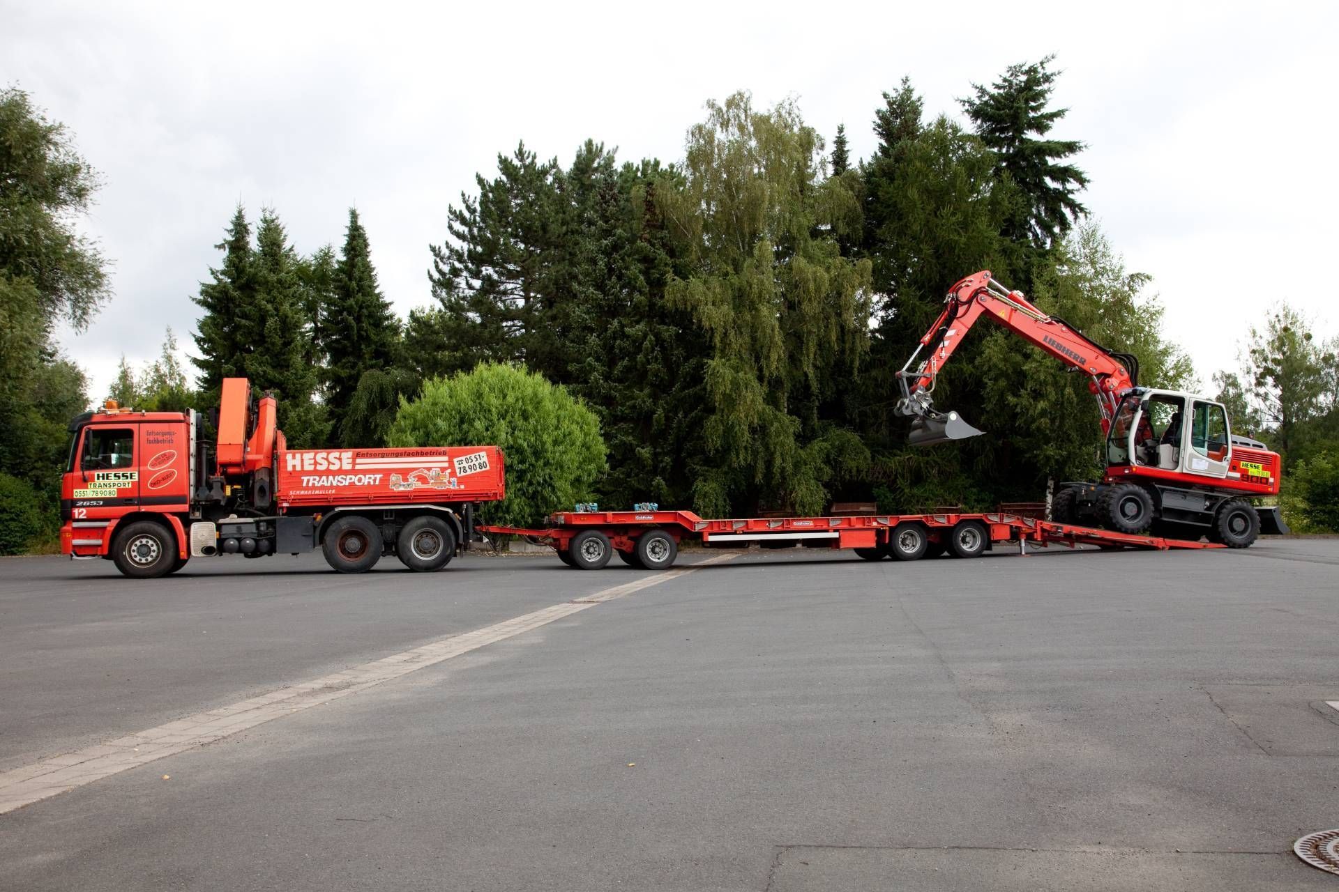 Ein roter Lastwagen zieht einen langen Tieflader mit einem orangefarbenen Bagger am Straßenrand.