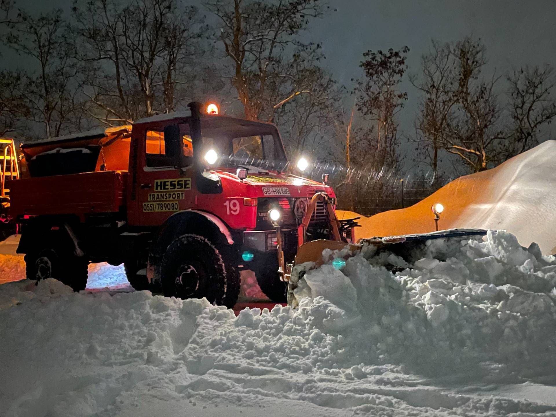 Ein roter Schneepflug räumt nachts tiefen Schnee neben einer großen Schneewehe