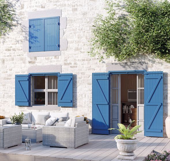 Maison en pierre avec volets bleus, patio aménagé avec mobilier en rotin : deux fauteuils, un canapé et une table basse. Décoration chaleureuse avec coussins et plantes vertes. Porte ouverte sur un intérieur avec bibliothèque. Végétation grimpante sur les murs et pots de fleurs autour du patio.