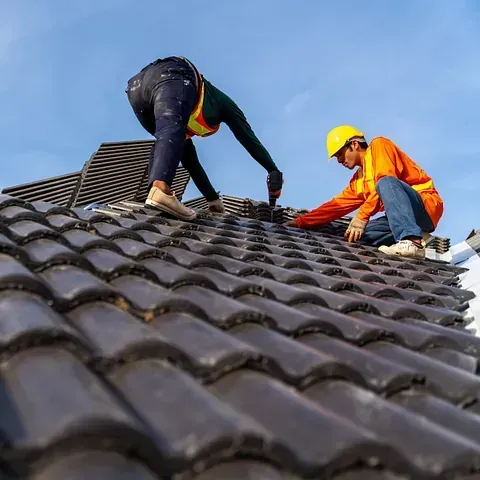 Dos trabajadores con cascos instalan tejas en un tejado empinado bajo un cielo azul.