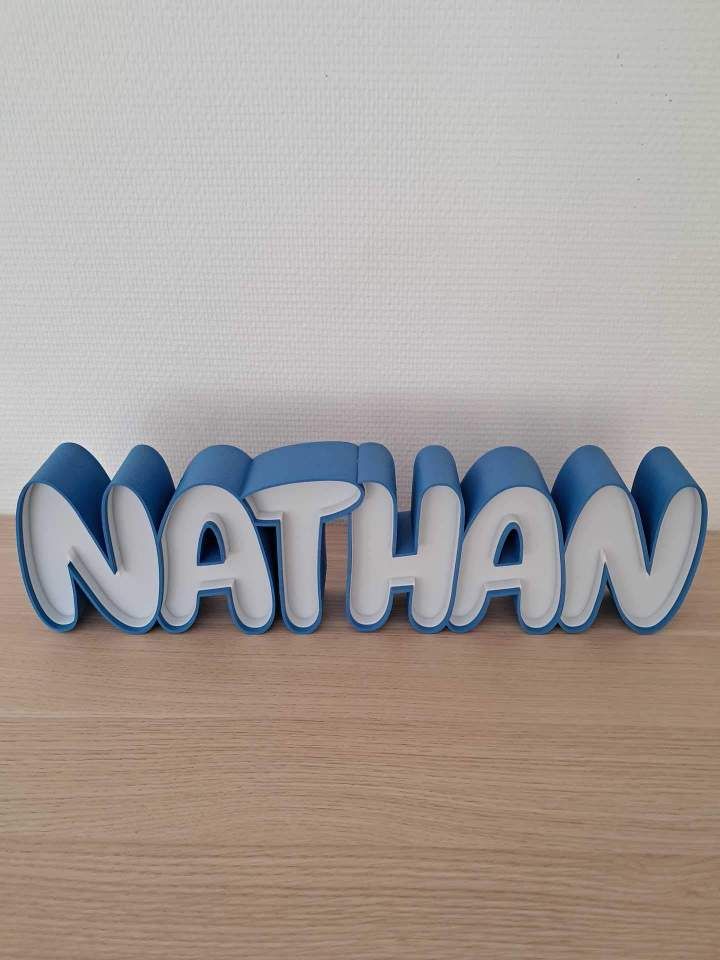 Inscription « NATHAN » bleue et blanche sur une surface en bois, devant un mur clair