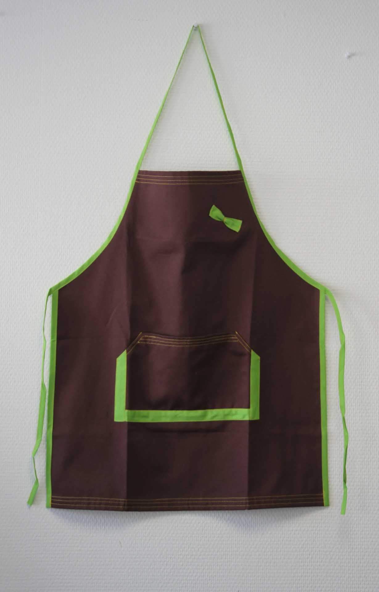 Tablier marron à bordure verte et poche avant, accroché au mur.