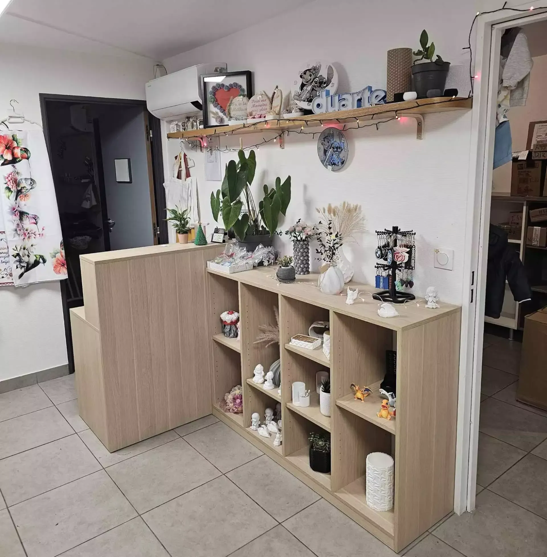 Petit comptoir de magasin avec étagères en bois clair, plantes en pot, décoration et sol carrelé dans un intérieur aux tons pastel