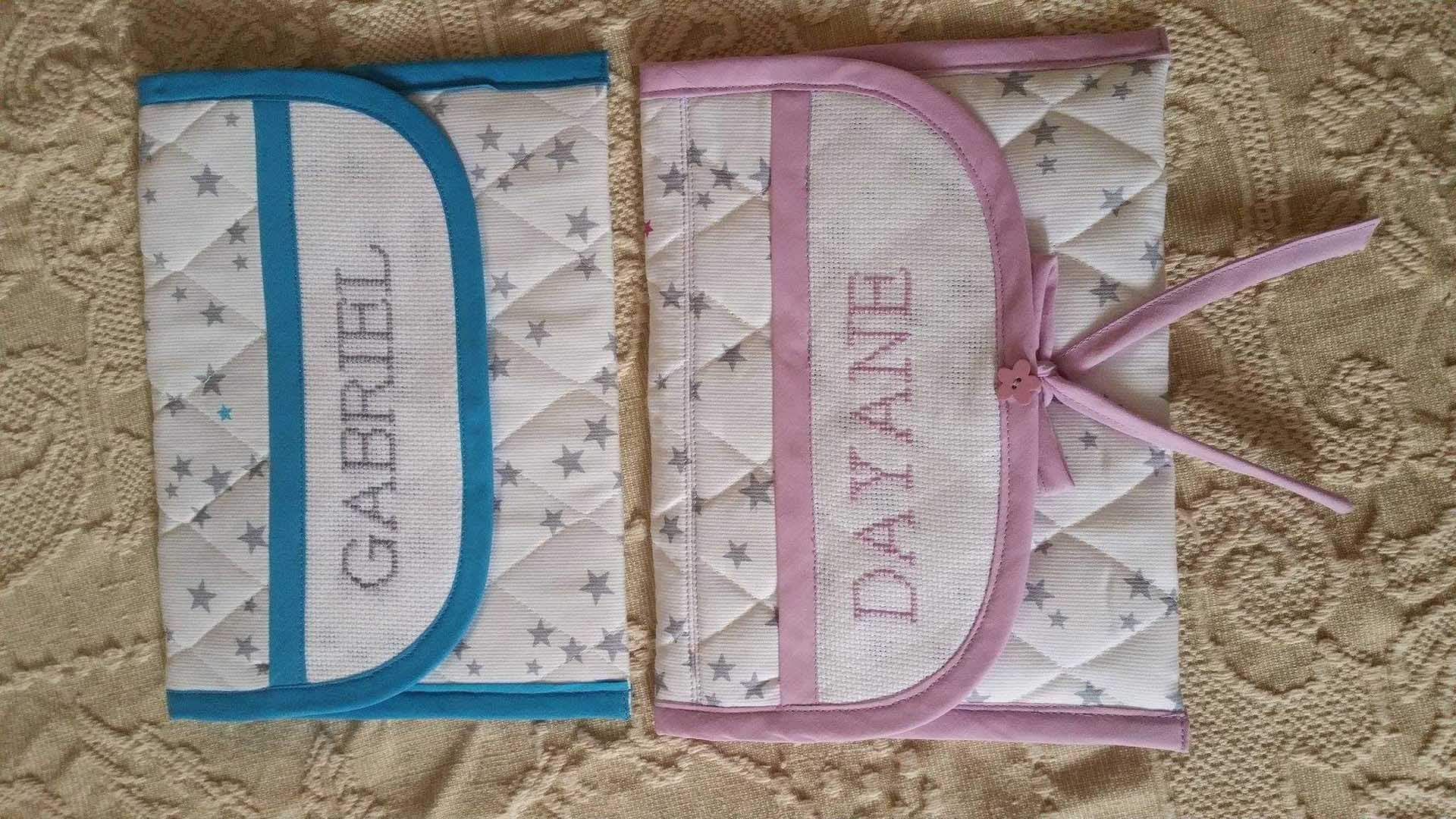 Deux pochettes matelassées zippées posées sur de la moquette, une bleue et une rose, toutes deux munies de poignées en ruban et de poches avant.