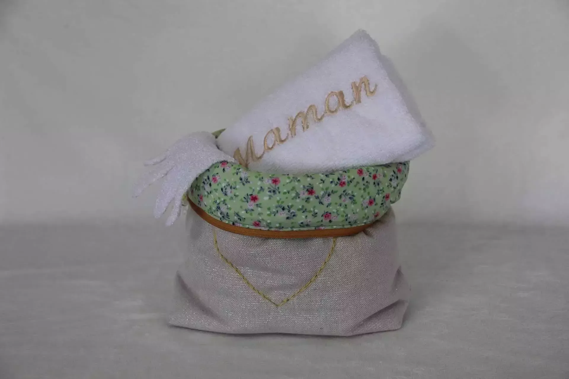 Pochette cadeau décorative en tissu blanc avec bande florale verte et étiquette nominative en écriture cursive sur fond uni