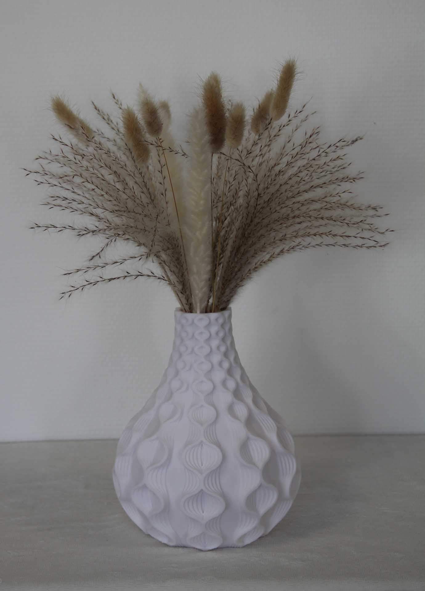 Vase blanc texturé contenant des tiges d'herbe beige séchées, posé sur une surface claire devant un mur uni.