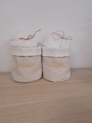 Deux paniers de rangement en tissu blanc, brodés de noms en or et munis de poignées en ficelle, posés sur une surface en bois.