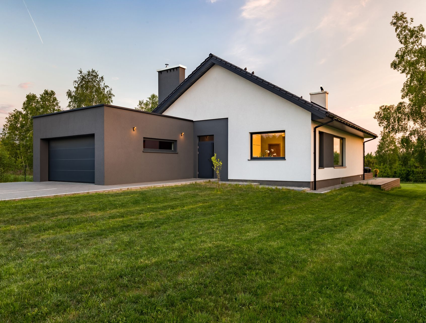 Une maison blanche moderne avec un garage gris se trouve sur une pelouse verte, des arbres en arrière-plan.