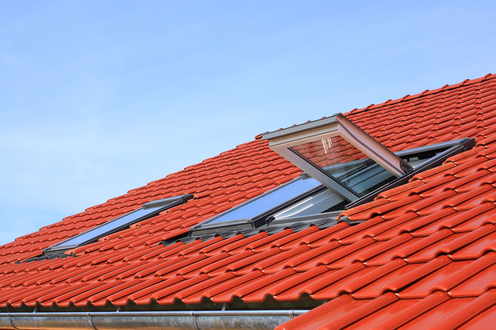 Toit en tuiles rouges avec deux puits de lumière ouverts contre un ciel bleu clair.