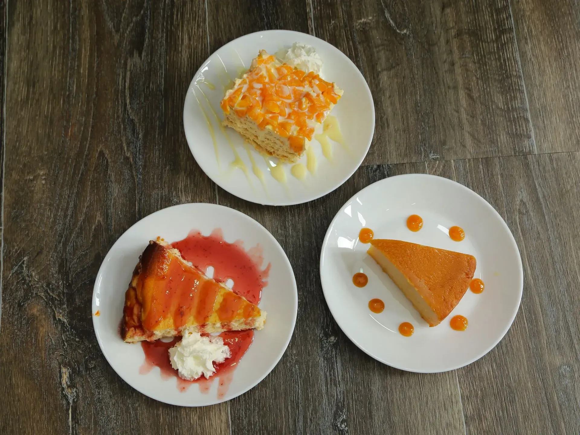 Tres rebanadas de pastel en platos blancos, rociadas con salsas y crema batida, dispuestas sobre una superficie de madera.