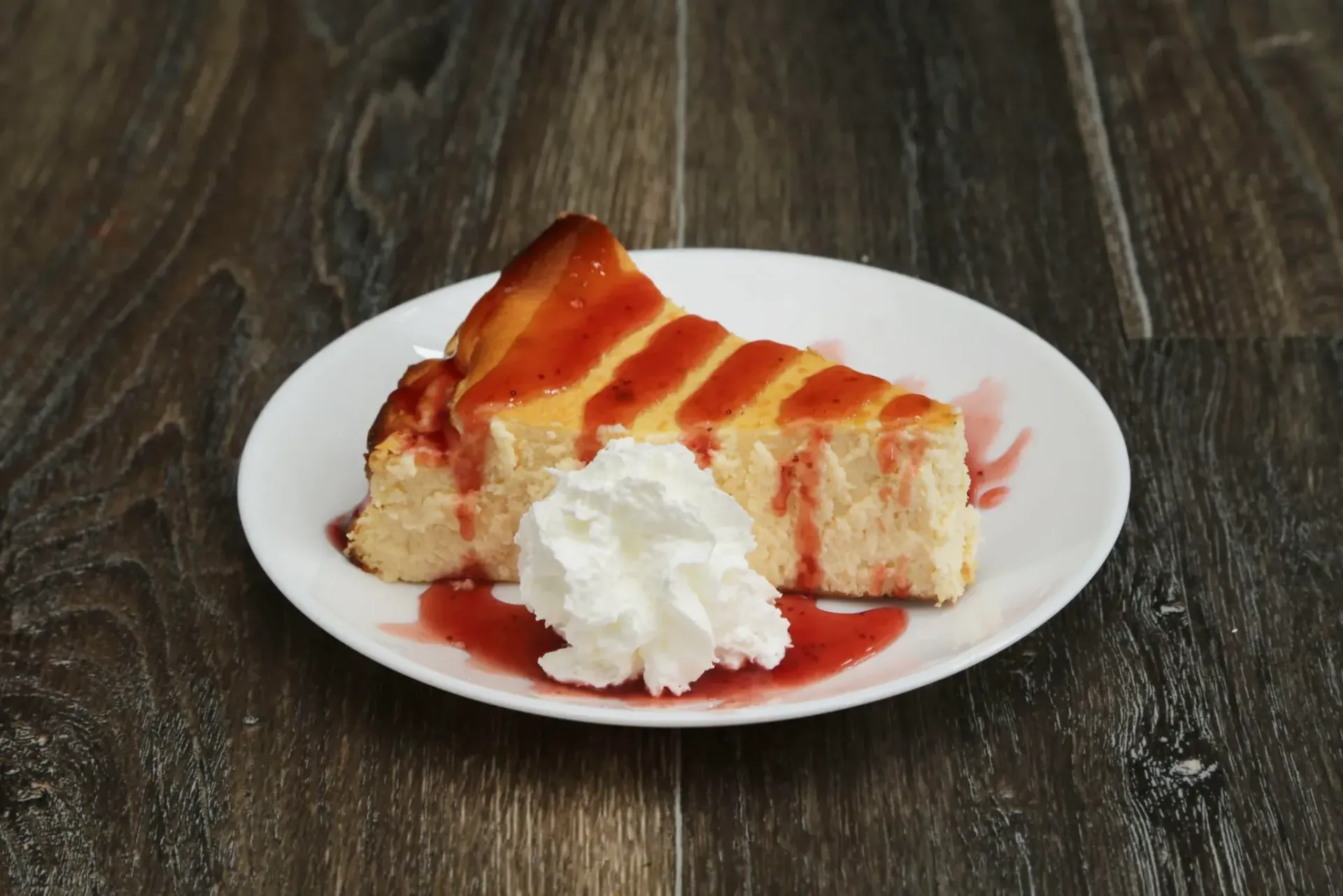 Porción de tarta de queso con salsa roja y nata montada en un plato blanco.
