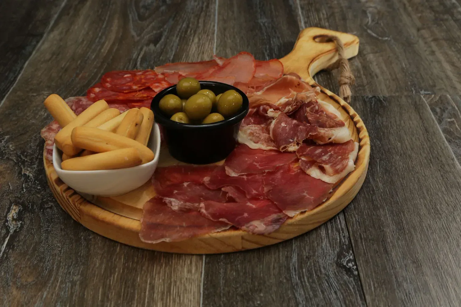 Tabla de embutidos: carnes curadas, aceitunas y palitos de pan en una tabla de madera.